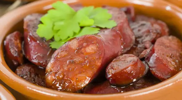 Slices of glistening chorizo al vino, simmered in a rich sauce, are served in a rustic clay bowl, garnished with fresh parsley.