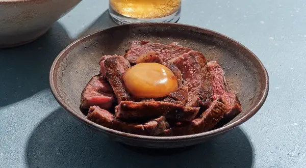 Sliced steak in a bowl topped with a glistening salt cured egg yolk, ready to be mixed into a rich, savory dish.