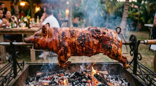 A whole spanish lechon, glistening and perfectly roasted, spins over glowing coals at an outdoor party.