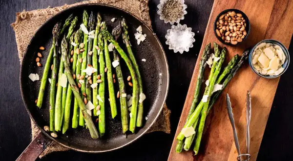 A pan of cooked asparagus with shaved parmesan and pine nuts, showing how to make asparagus a delicious side dish.
