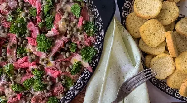Thinly sliced beef carpaccio, adorned with capers and parsley, is served with toasted bread rounds.
