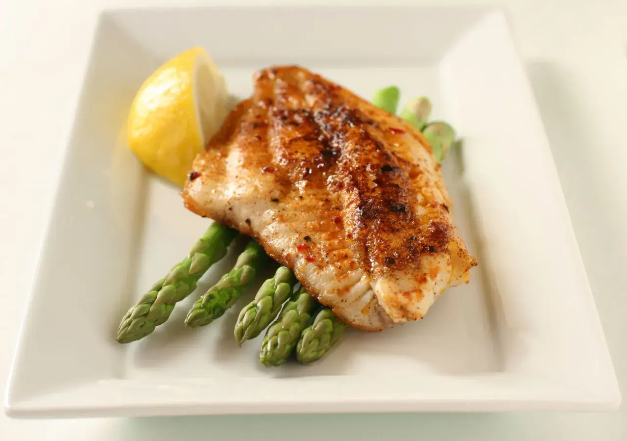 A perfectly seasoned fish fillet rests on a bed of asparagus, a lemon wedge on the side. A delightful dish inspired by Spanish fish recipes.