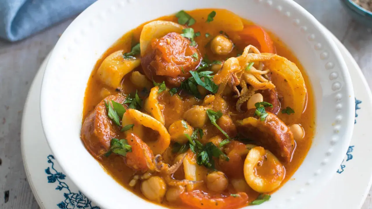A hearty Spanish stew with tender squid rings, chickpeas, and chorizo in a rich tomato sauce, garnished with fresh parsley.