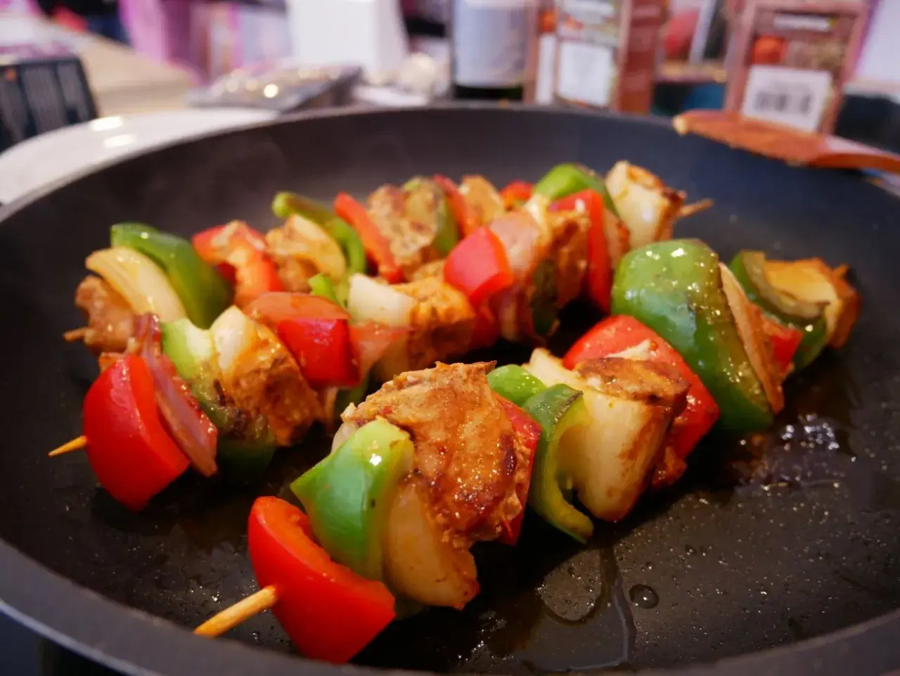 Pinchos morunos sizzling in a pan, with chunks of marinated meat, red and green bell peppers, and onions on wooden skewers.