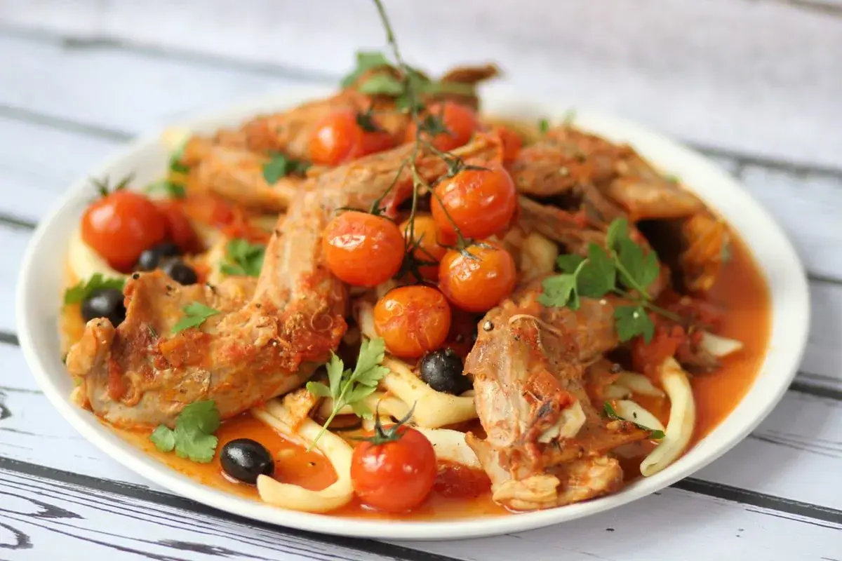 A hearty rabbit stew with tender meat, cherry tomatoes, olives, and pasta, garnished with fresh parsley.