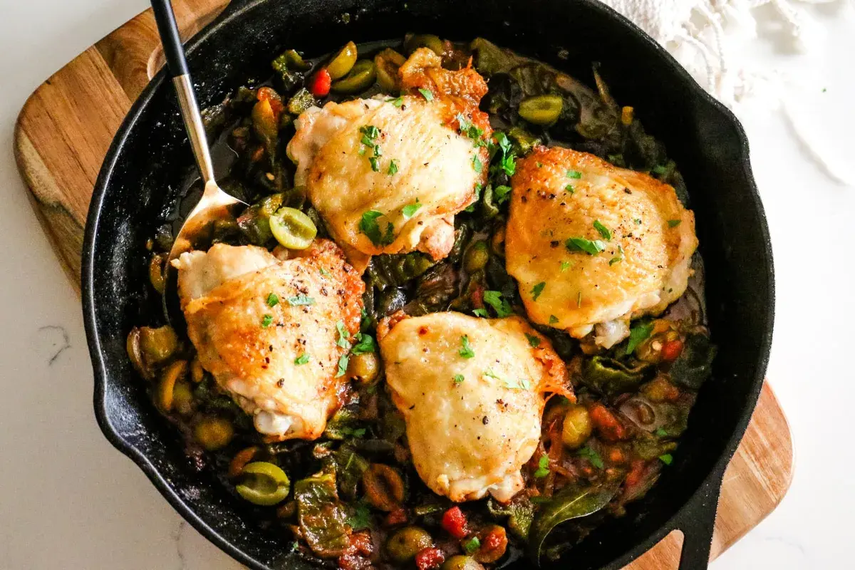 Four golden chicken thighs nestled in a rich, rustic chicken basque stew with olives and peppers, garnished with fresh parsley.
