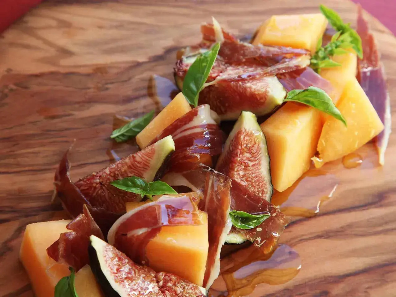 A refreshing melon con jamon appetizer with fresh figs and basil, drizzled with honey on a wooden board.