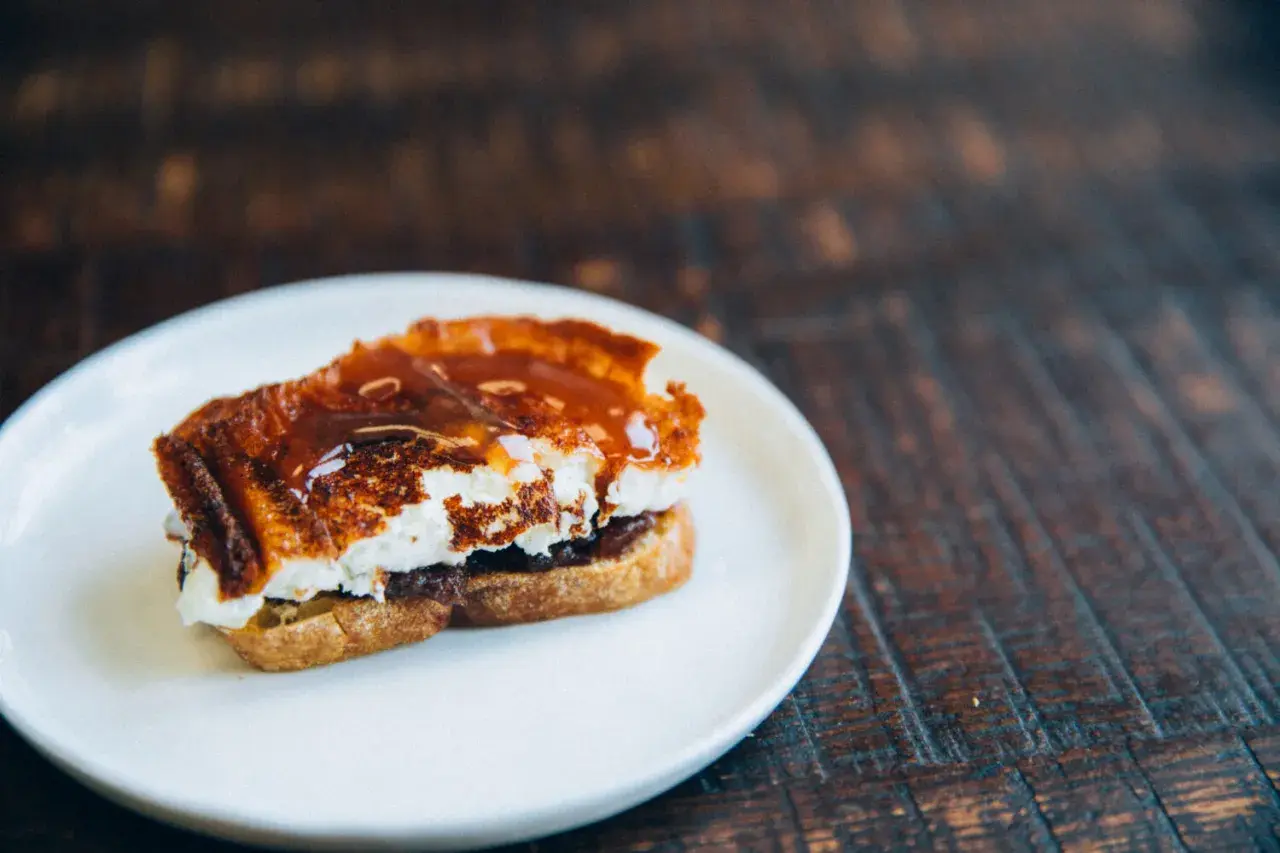 A decadent goat cheese toast, caramelized and drizzled with a rich, glossy sauce, sits on a white plate.
