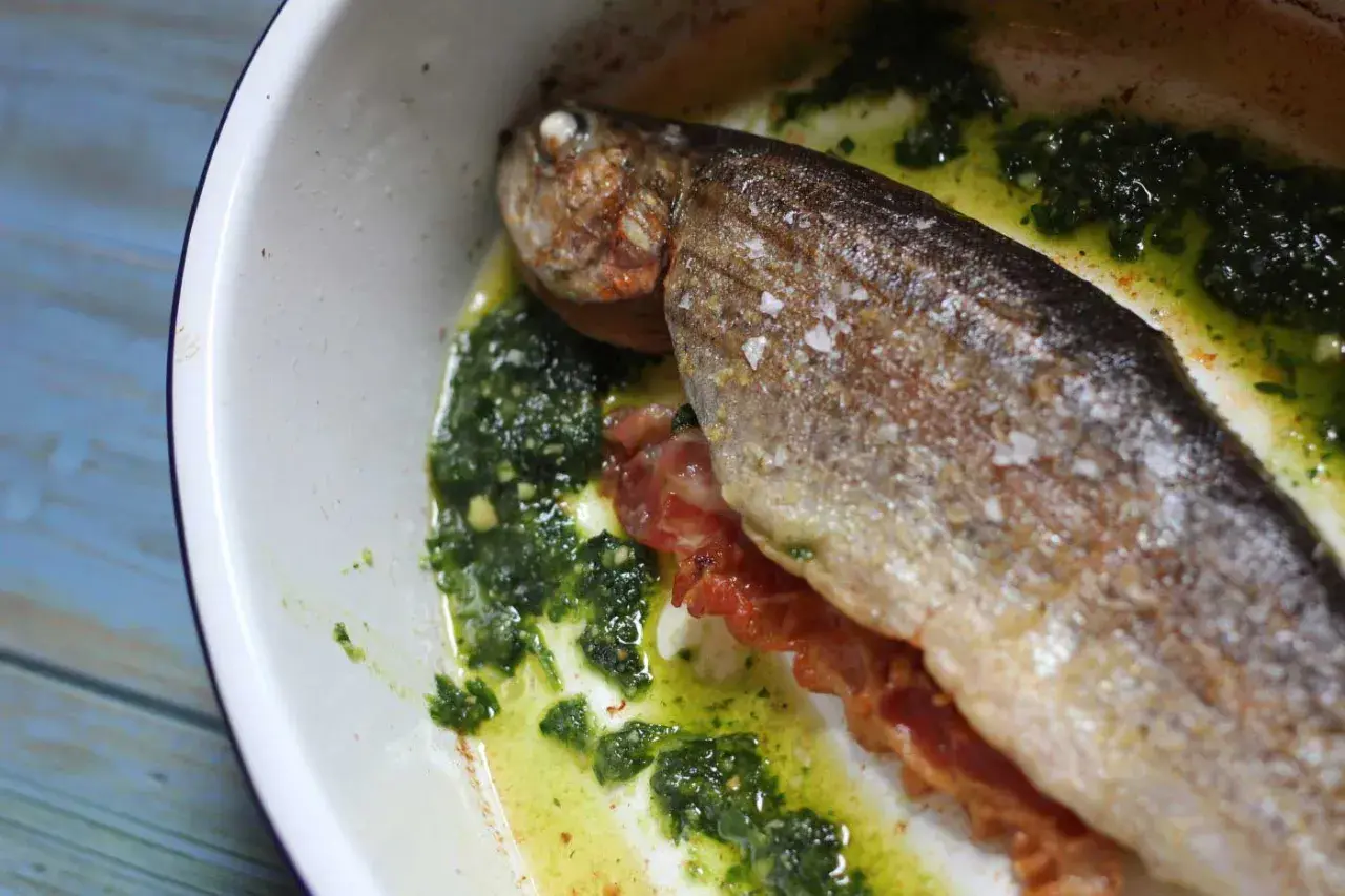 A whole la trucha, seasoned with salt, rests in a white dish with a vibrant green herb sauce and crispy bacon.