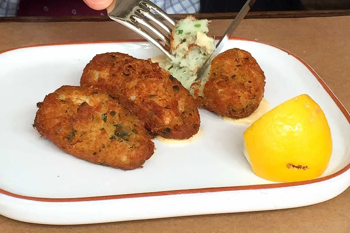 Golden brown cod fritters served with a dollop of mashed potatoes and a lemon wedge. A fork is lifting a bite of the fritter.
