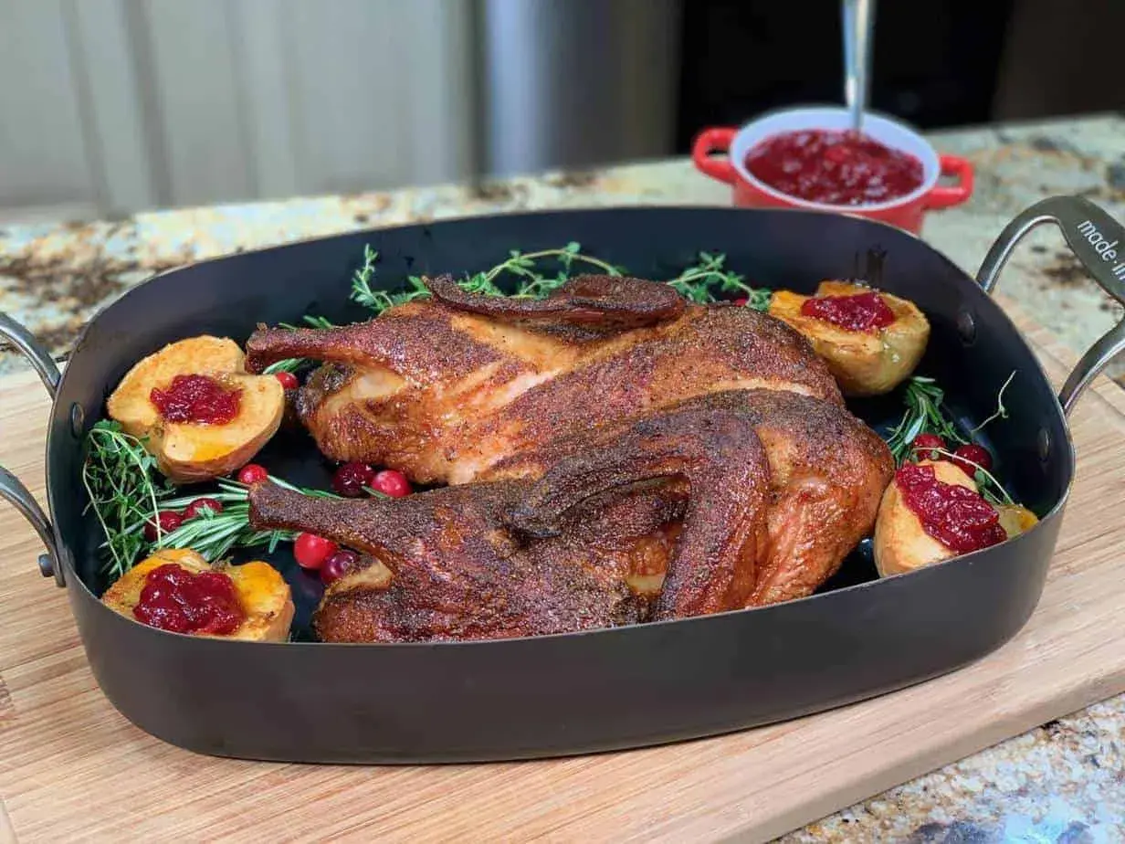 A beautifully roasted smoked duck rests in a dark roasting pan, garnished with fresh rosemary, cranberries, and baked apple halves topped with cranberry sauce.