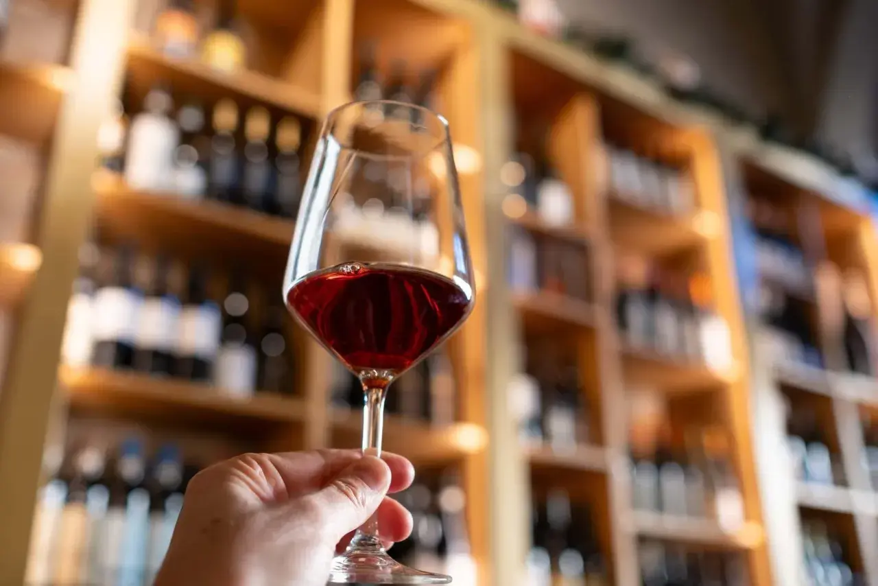 A hand holds a glass of red wine, with shelves of wine bottles in the background. Some believe certain red wines may boost metabolism.