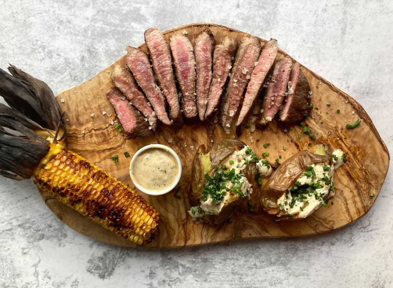 Juicy pork presa, grilled corn, and loaded baked potatoes served on a wooden board.