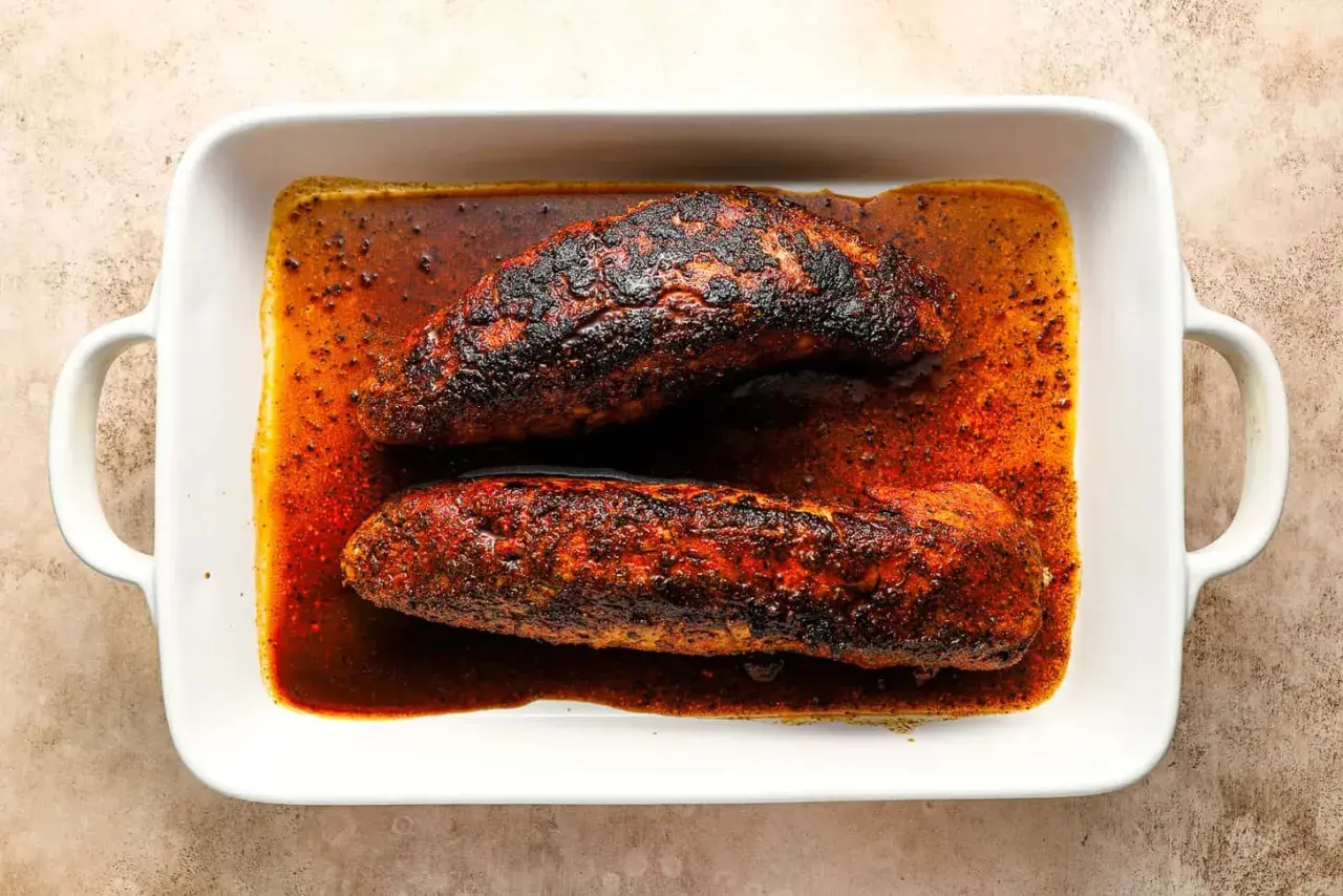 Two seasoned pork tenderloins, ready for a delicious solomillo recipe, rest in a white baking dish filled with rich, spiced juices.