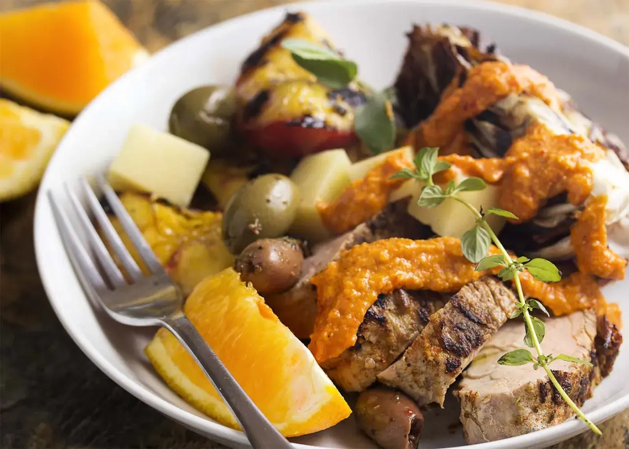 Slices of grilled spanish pork tenderloin with olives, orange wedges, and a vibrant romesco sauce.