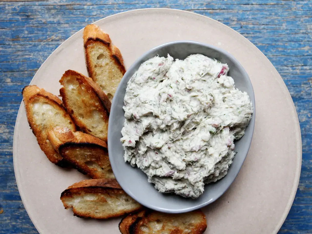 A bowl of creamy bluefish pate with herbs and red onion, served with toasted baguette slices on a light-colored plate.