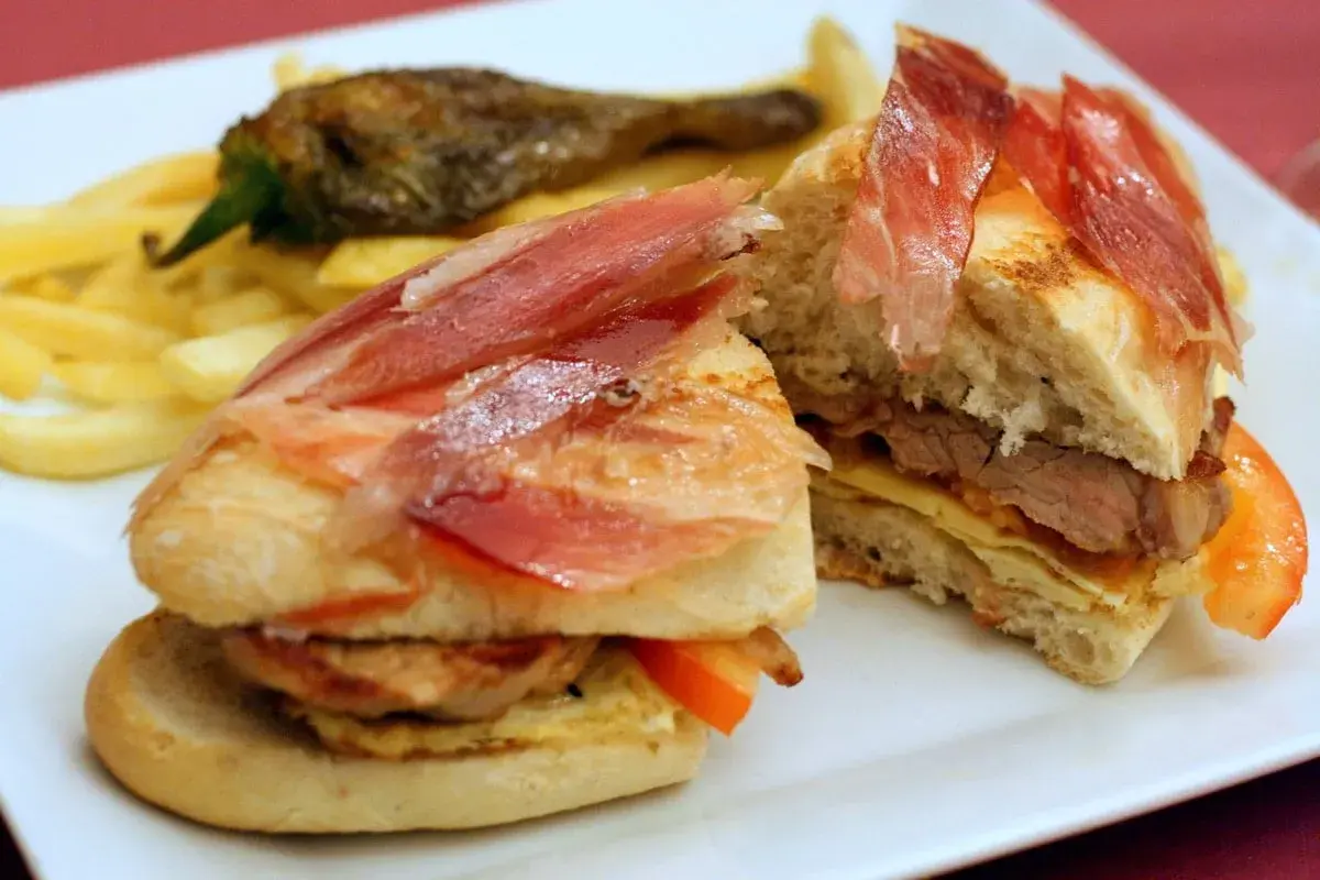 A delicious bocadillo with tender meat, cured ham, and fresh tomato, served with crispy fries and a roasted pepper.