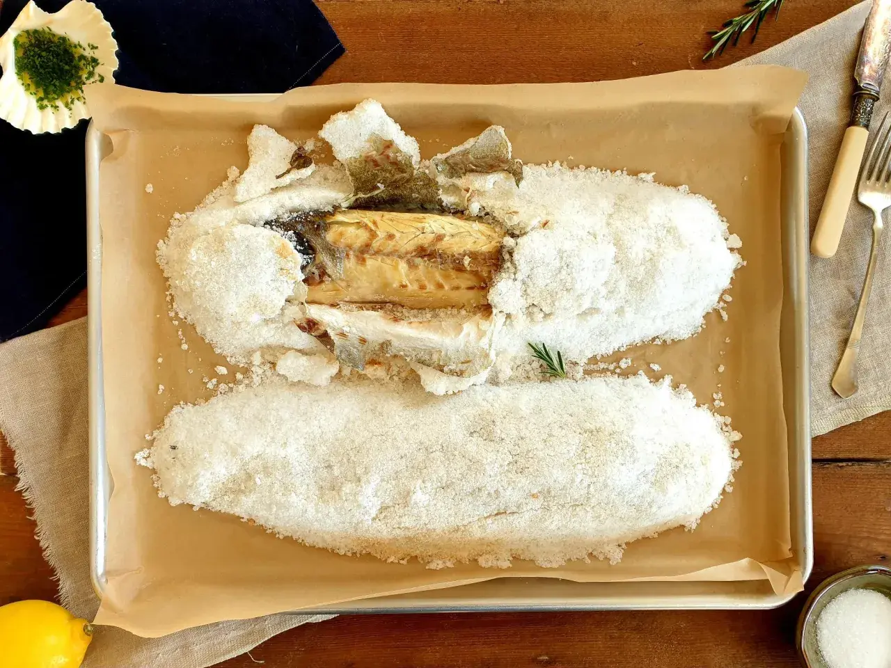 A whole salt crusted sea bass, baked to perfection, rests on a baking sheet. A lemon and herbs complete the dish.
