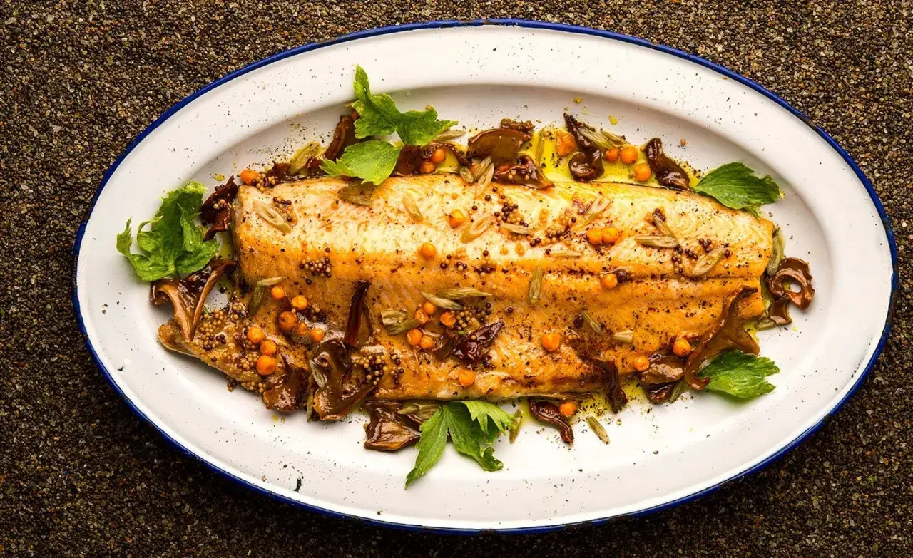 A beautifully broiled trout, glistening with olive oil and garnished with mustard seeds, sea buckthorn berries, and fresh herbs on a white enamel platter.