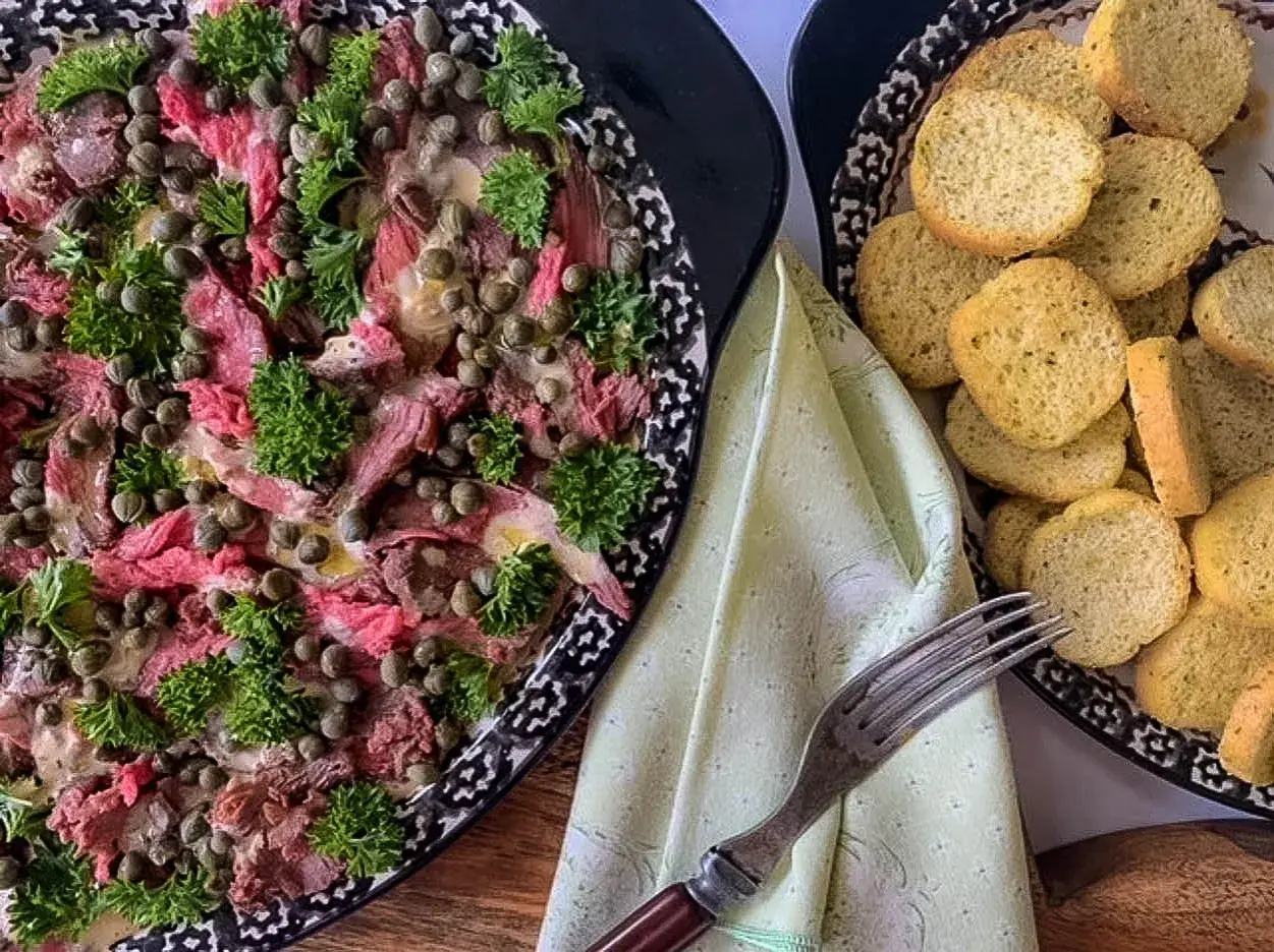 Thinly sliced beef carpaccio, adorned with capers and parsley, is served with toasted bread rounds.