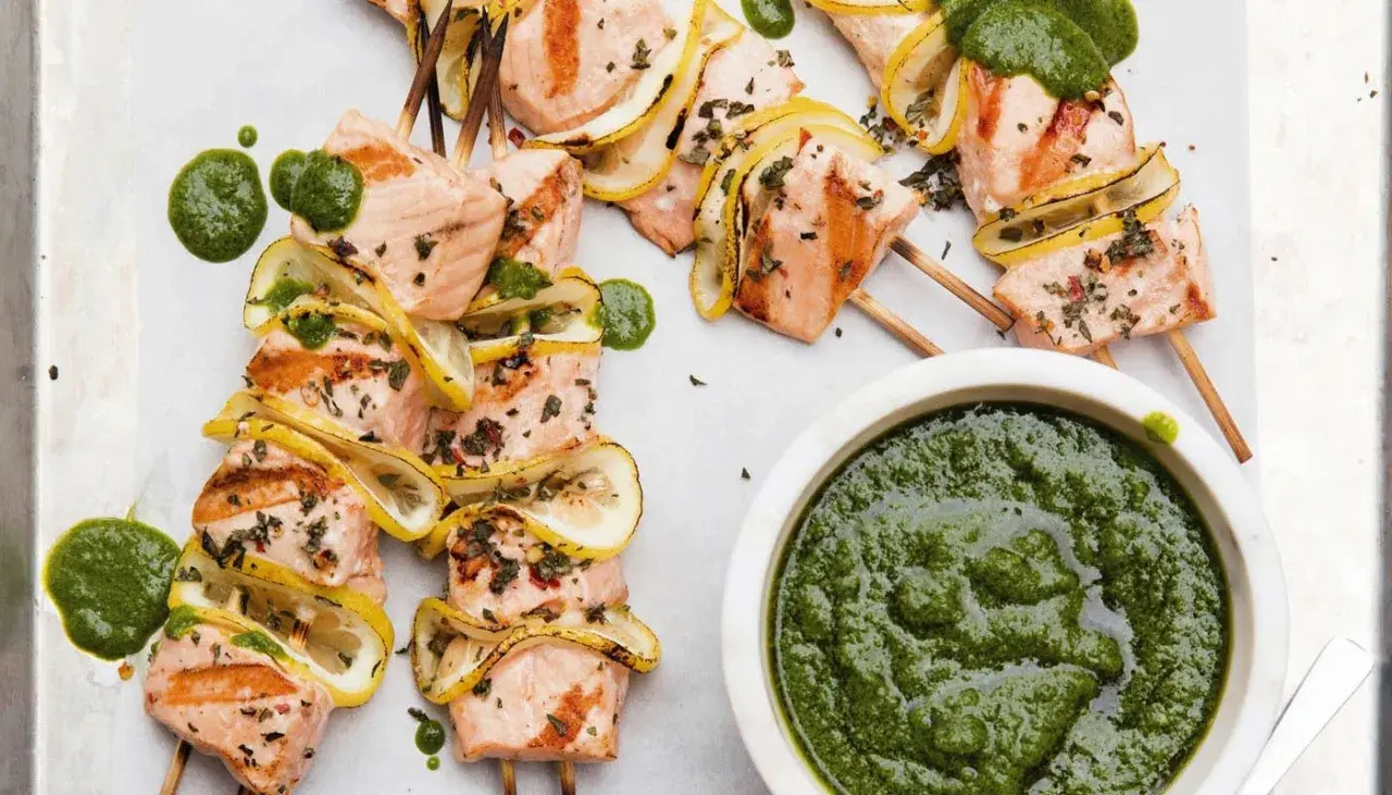 Grilled salmon skewers with lemon slices and a vibrant green sauce, ready to be dipped.