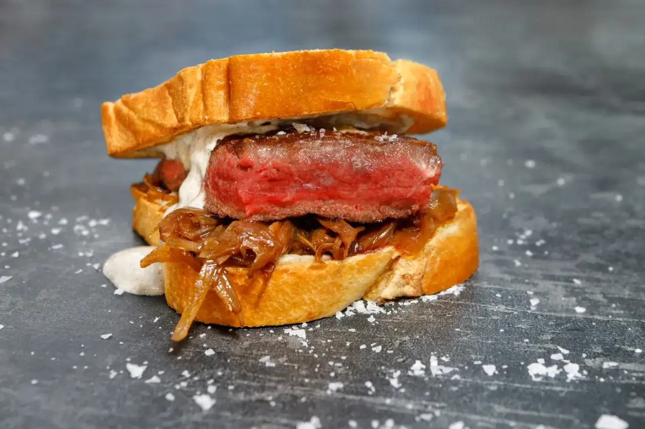 A juicy pepito de ternera, a tender steak sandwich on toasted bread with caramelized onions and a creamy sauce, sprinkled with sea salt.