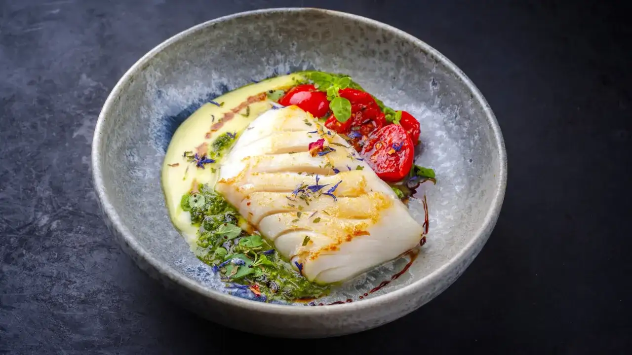 A beautifully plated poached fish recipe, featuring a white fish fillet with grilled marks, served with creamy sauce, pesto, and cherry tomatoes.