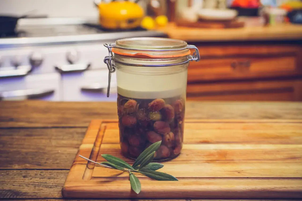 Jar of olives in brine, ready for curing. A fresh olive branch rests on the wooden cutting board.