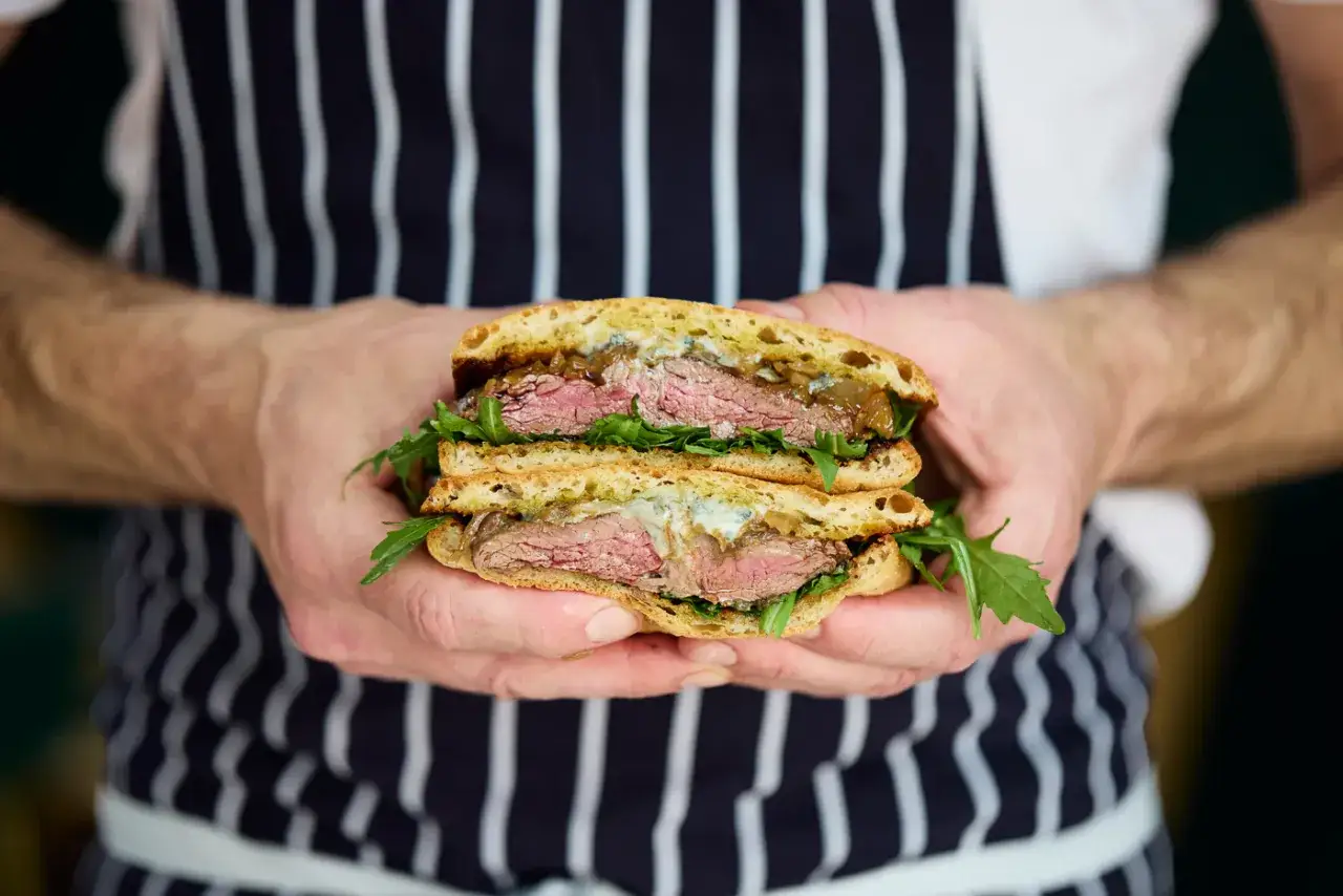 A chef holds a delicious pepito sandwich, overflowing with tender steak, caramelized onions, and fresh greens.