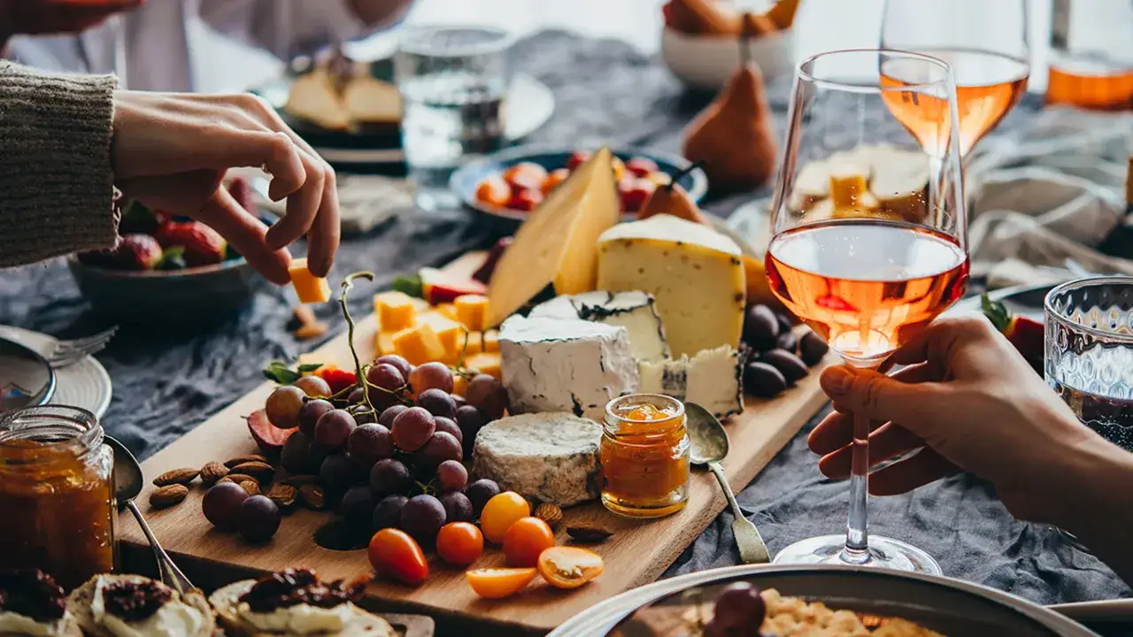 A cheese board with grapes, nuts, and various types of wine, including ros&eacute;, is being enjoyed by friends.