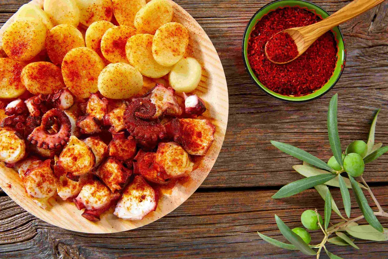 A delicious plate of pulpo a feira, tender octopus seasoned with paprika, served with boiled potatoes. A small bowl of paprika and olives complete the rustic presentation.