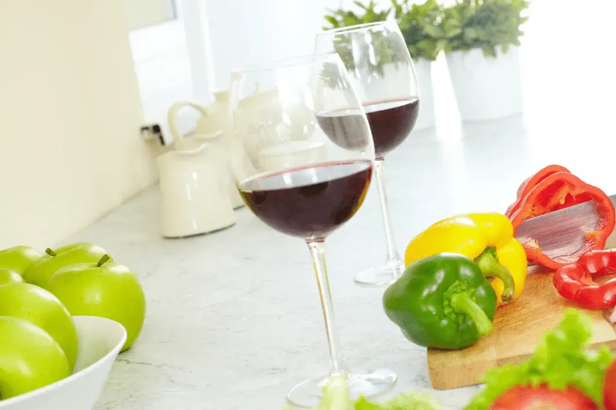 Two glasses of Carmenere wine sit on a kitchen counter next to fresh apples and colorful bell peppers, ready for a meal.