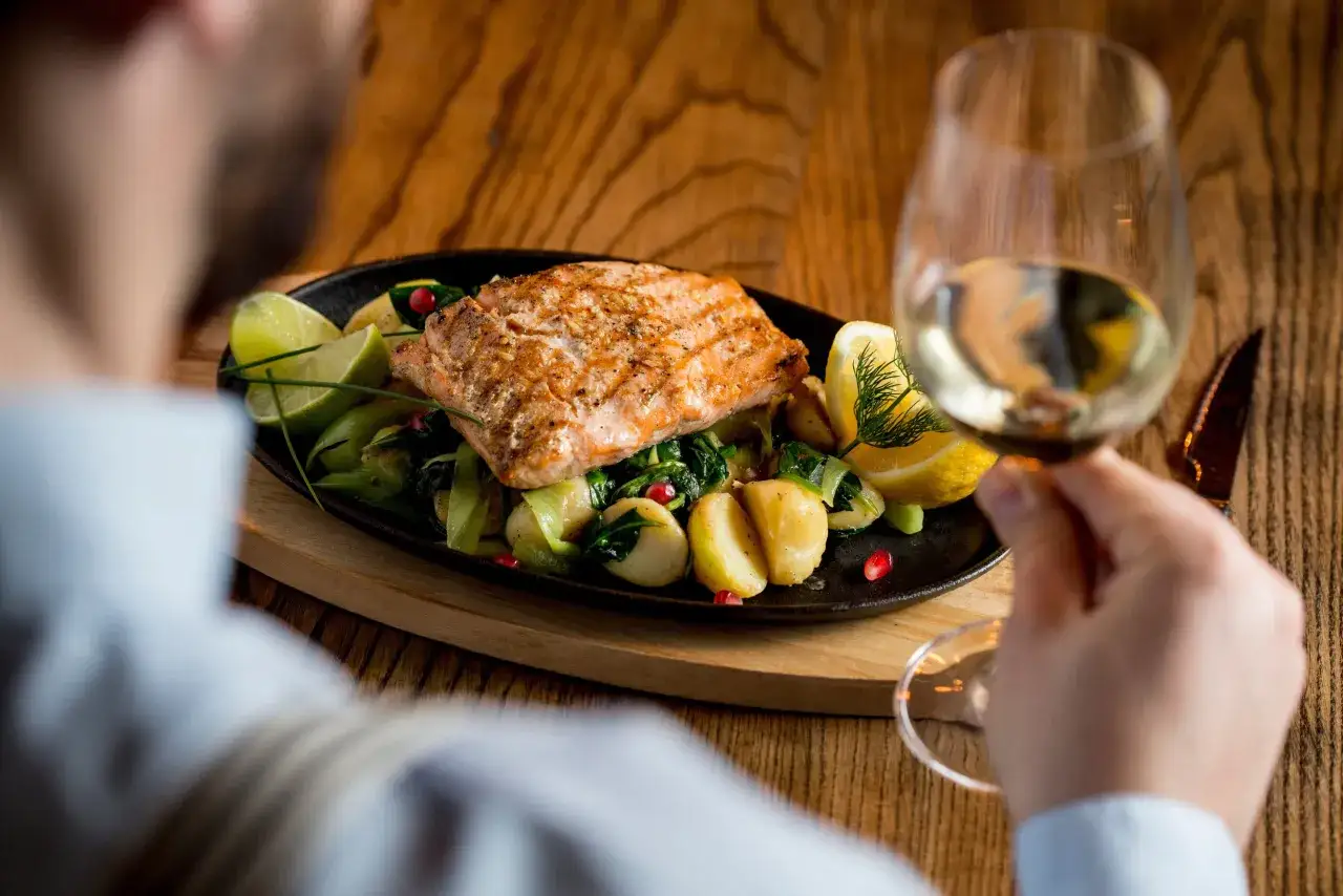 A man enjoys a perfectly cooked salmon dish with potatoes, greens, and lemon, paired with a glass of white wine, anticipating the crisp pinot grigio taste.