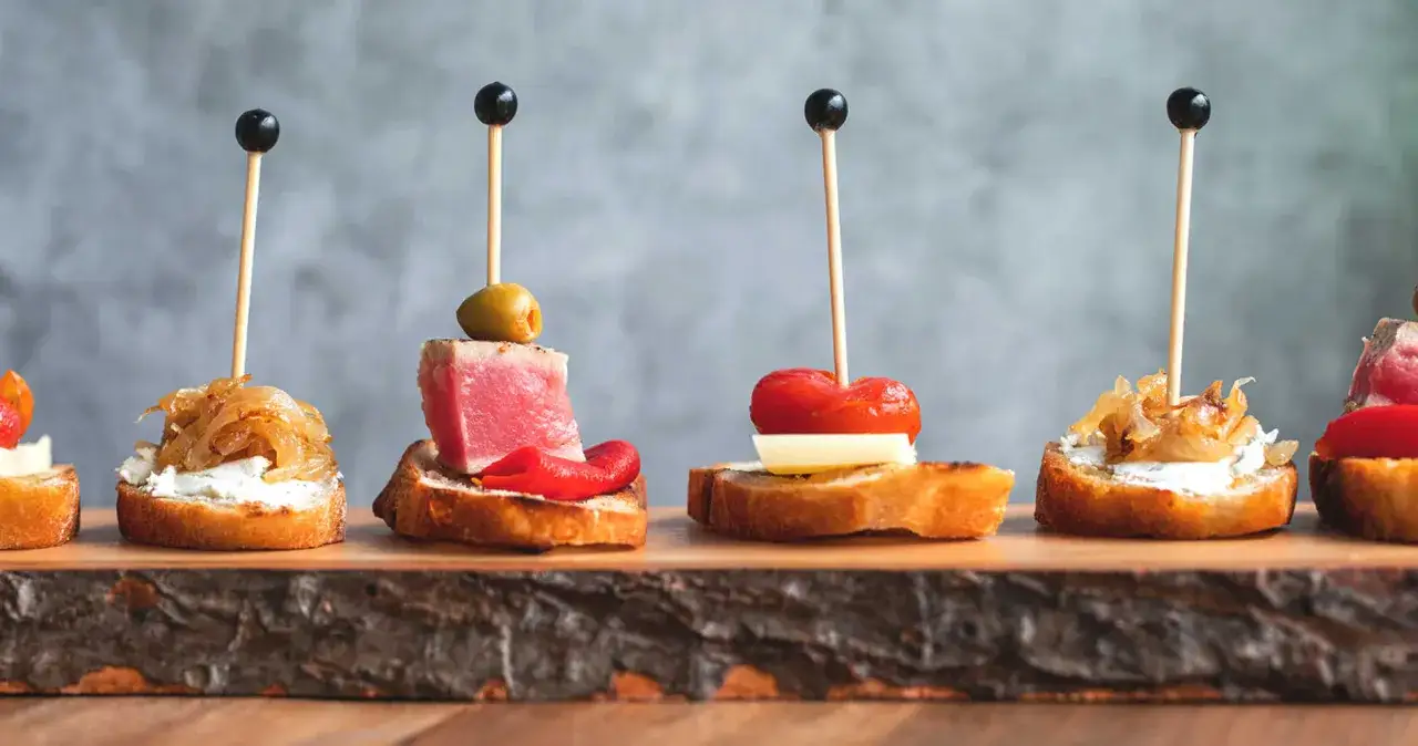 A beautiful charcuterie board featuring various tapas on toasted bread, including tuna, caramelized onions, and roasted peppers, all artfully arranged.