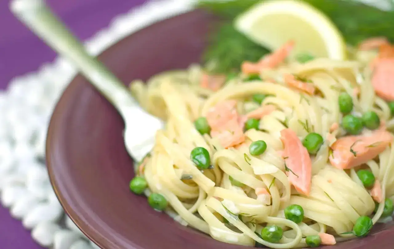 A delicious plate of linguine with flakes of smoked salmon, bright green peas, and fresh dill, garnished with a lemon wedge.