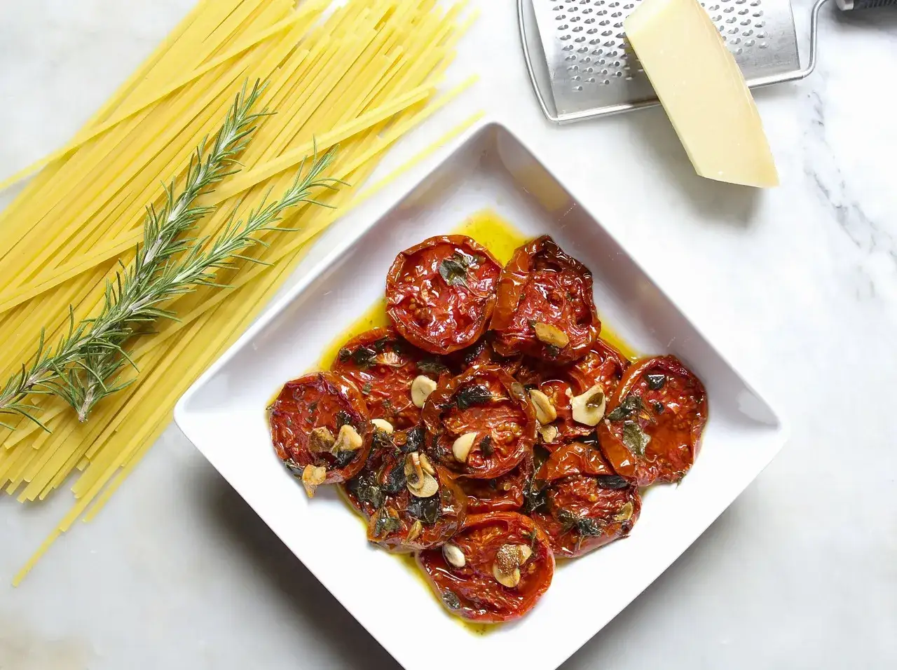 A white dish holds glistening slow roasted tomatoes with garlic and herbs, next to a bundle of spaghetti, a grater, and a wedge of Parmesan cheese.