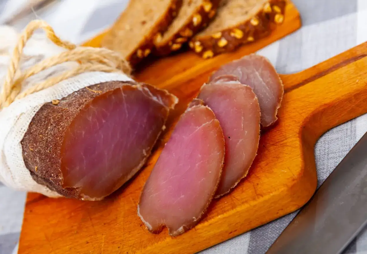 A whole and sliced cured pork loin, wrapped in cheesecloth and twine, sits on a wooden board with slices of rustic bread.