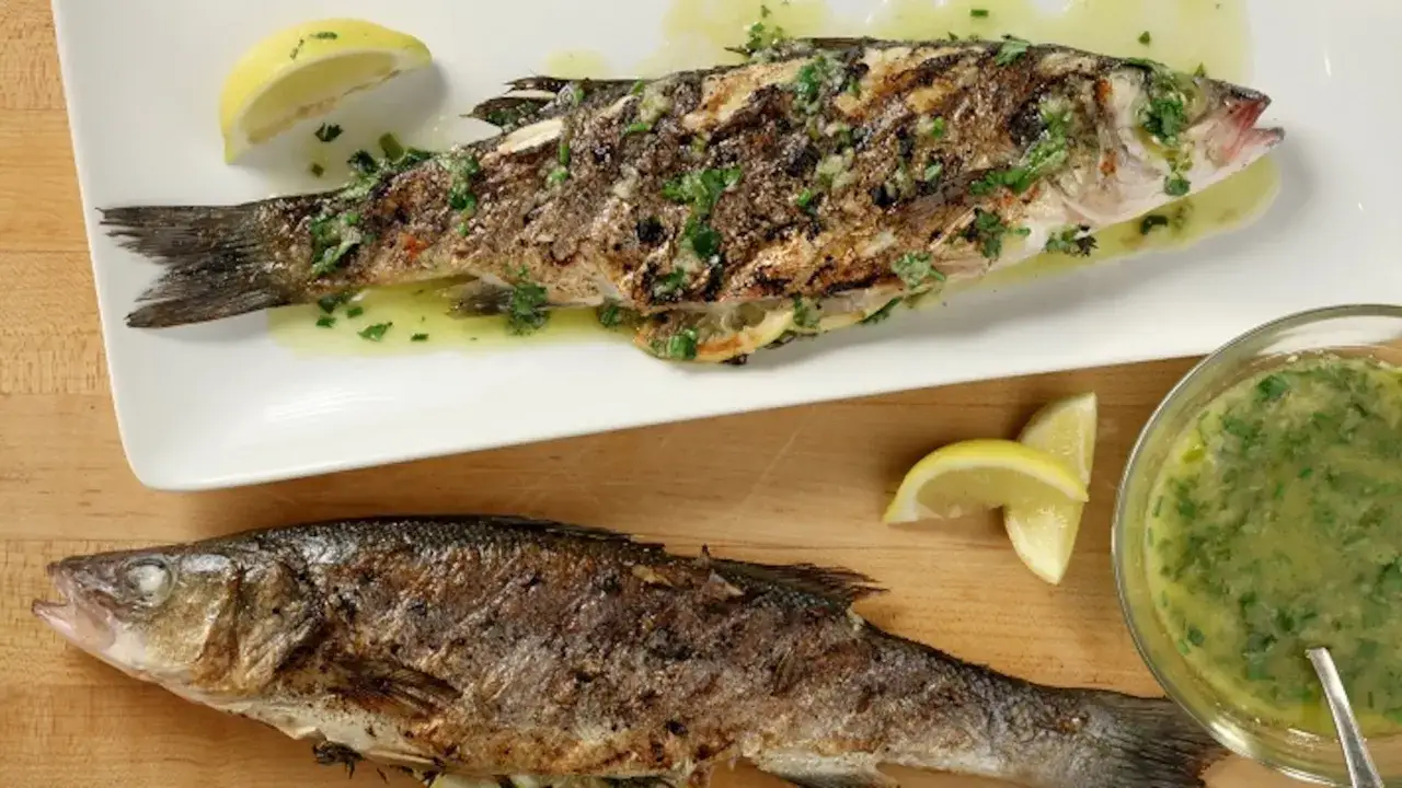 Two whole grilled fish, seasoned with herbs and lemon, sit on a white platter and wooden board, ready to be enjoyed.