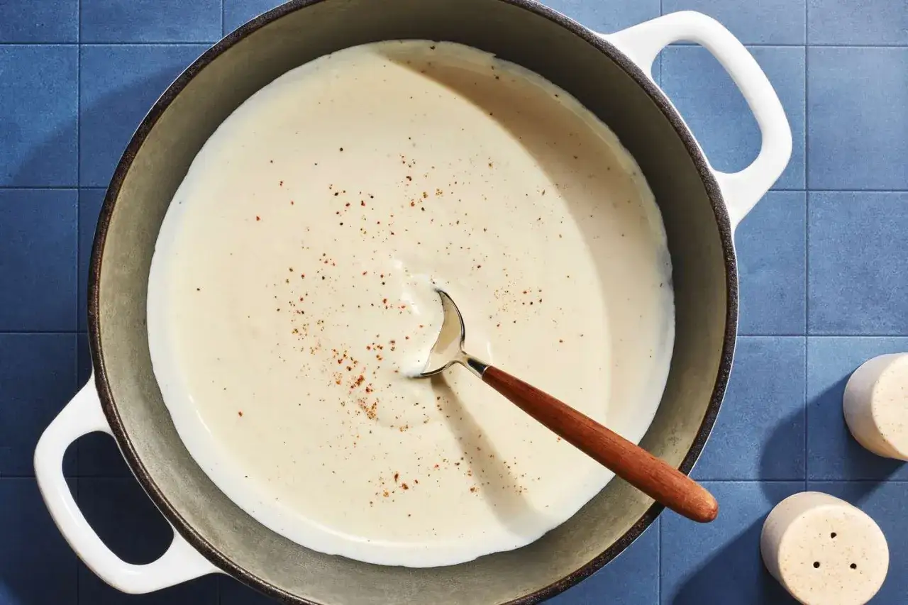 A creamy sauce, likely cr&egrave;me fra&icirc;che, simmers in a pot, seasoned with pepper. A wooden-handled spoon rests in the rich mixture.