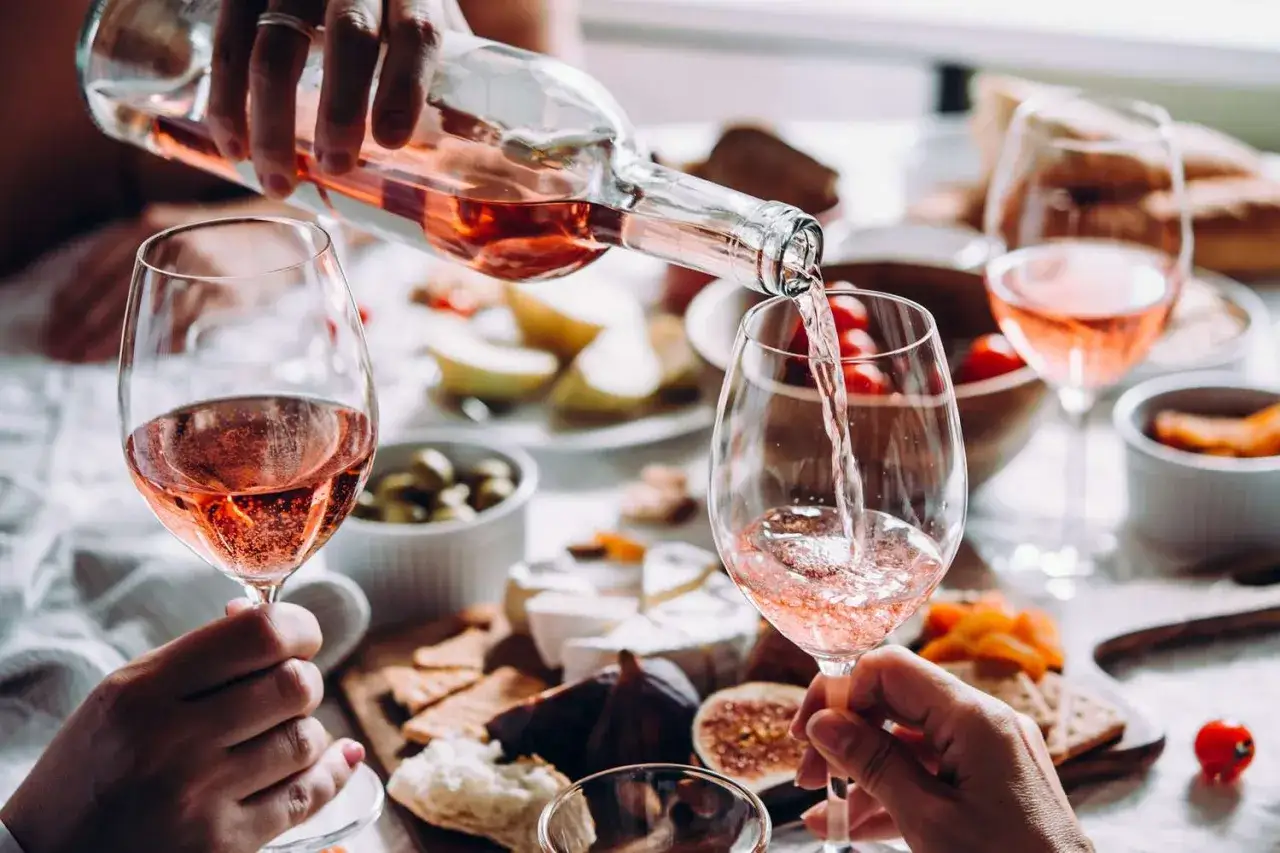 Pouring ros&eacute; wine, a delightful choice for a healthy indulgence, into a glass. A spread of cheese, crackers, and fruit suggests a perfect pairing.