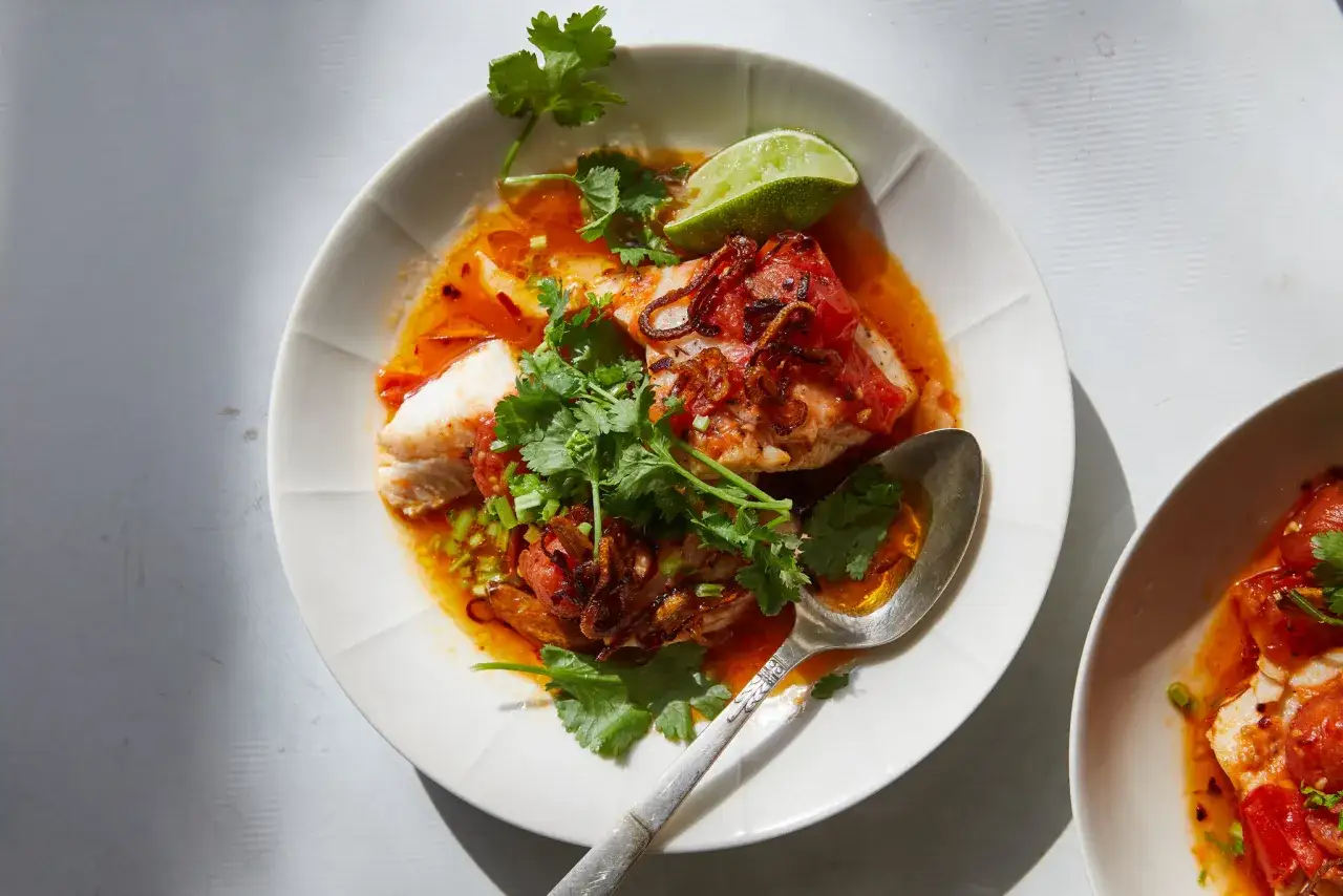 A vibrant dish of poached fish in a rich tomato broth, garnished with fresh cilantro, crispy fried onions, and a lime wedge. This looks like a delicious recipe for poached fish.