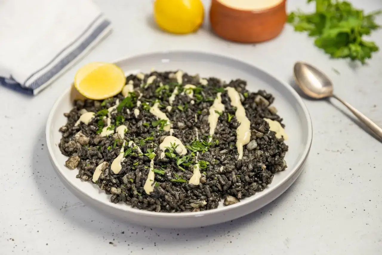 A delicious plate of squid ink rice, drizzled with a creamy sauce and garnished with fresh parsley. A lemon wedge sits on the side, ready to add a zesty finish.