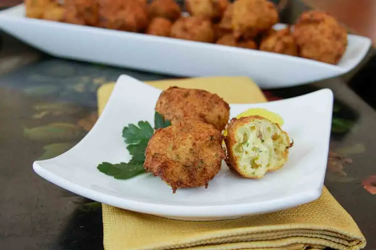 Golden brown cod fritters, one cut open to reveal a fluffy interior with herbs, served on a white plate with parsley.