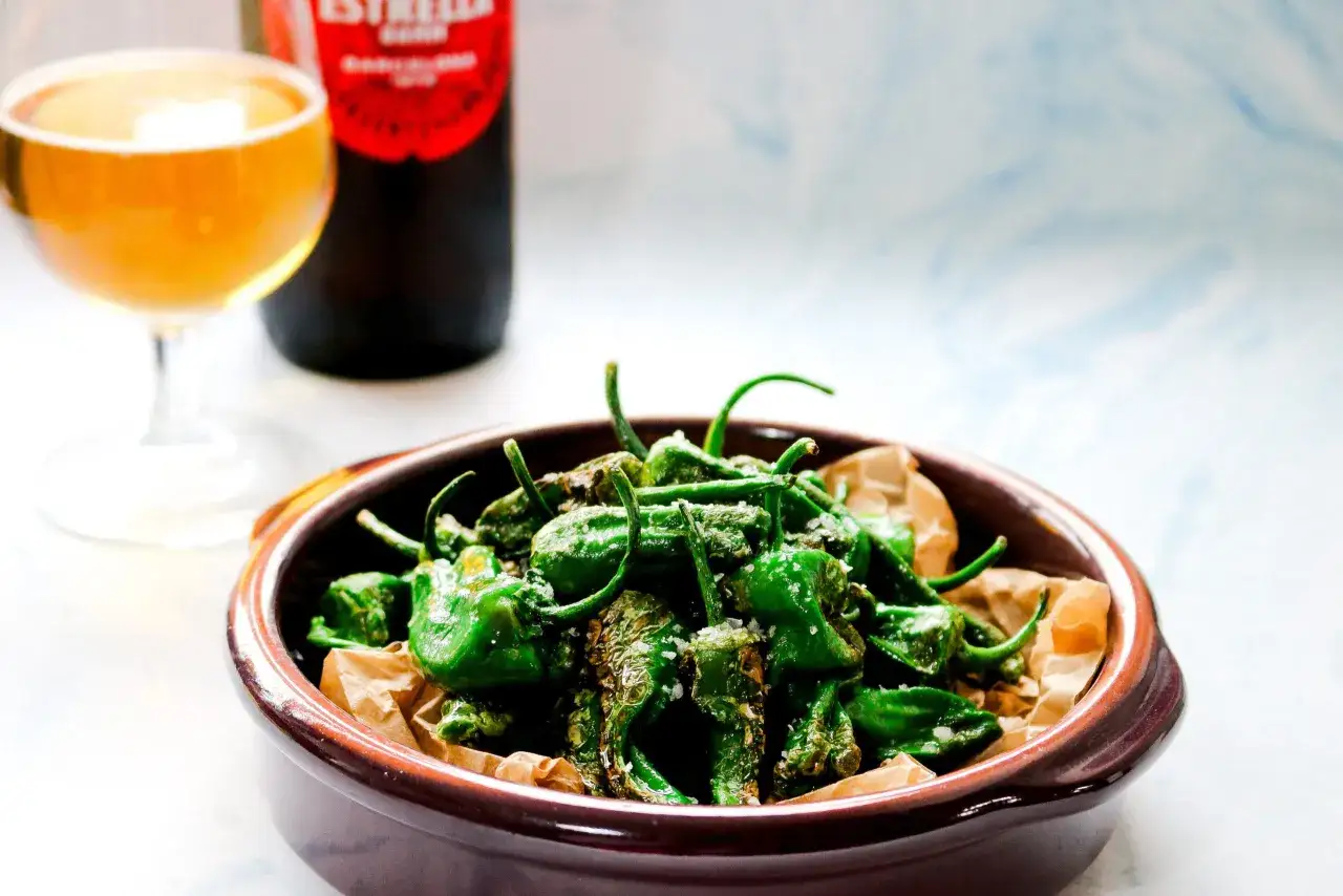 A bowl of perfectly fried padron peppers, lightly salted, sits next to a glass of beer and a bottle of Estrella.