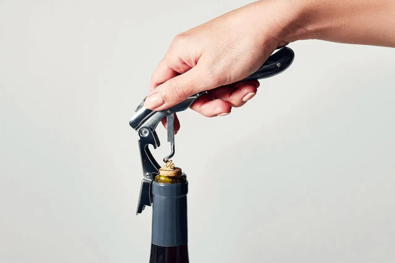 A hand uses a waiter's corkscrew to demonstrate how to open a wine bottle, with the worm just starting to enter the cork.