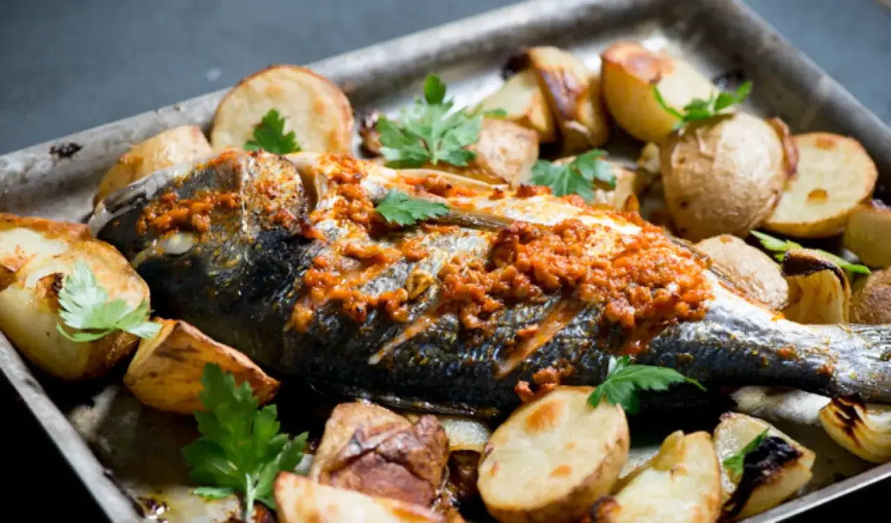 A whole sea bream, baked with roasted potatoes and garnished with fresh parsley, is presented on a metal tray.