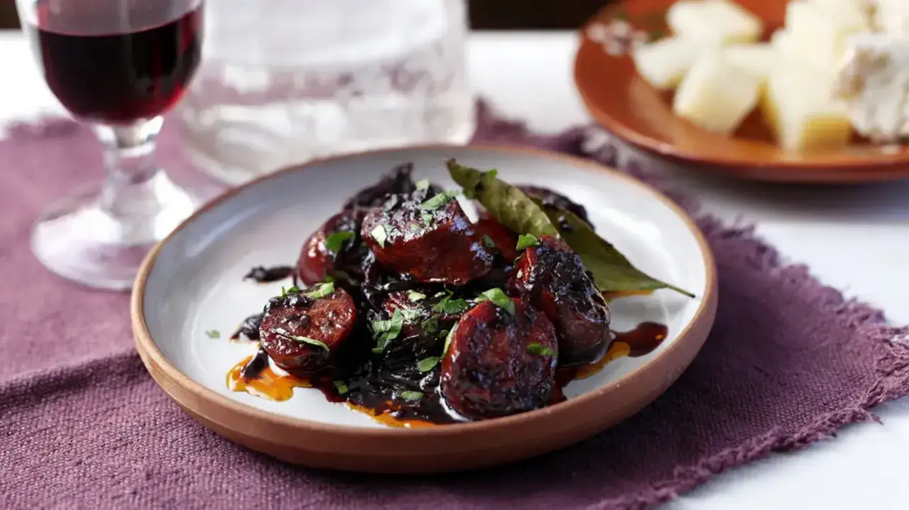 A plate of rich chorizo al vino, glistening with a dark sauce and garnished with fresh parsley. A glass of red wine and cheese are in the background.