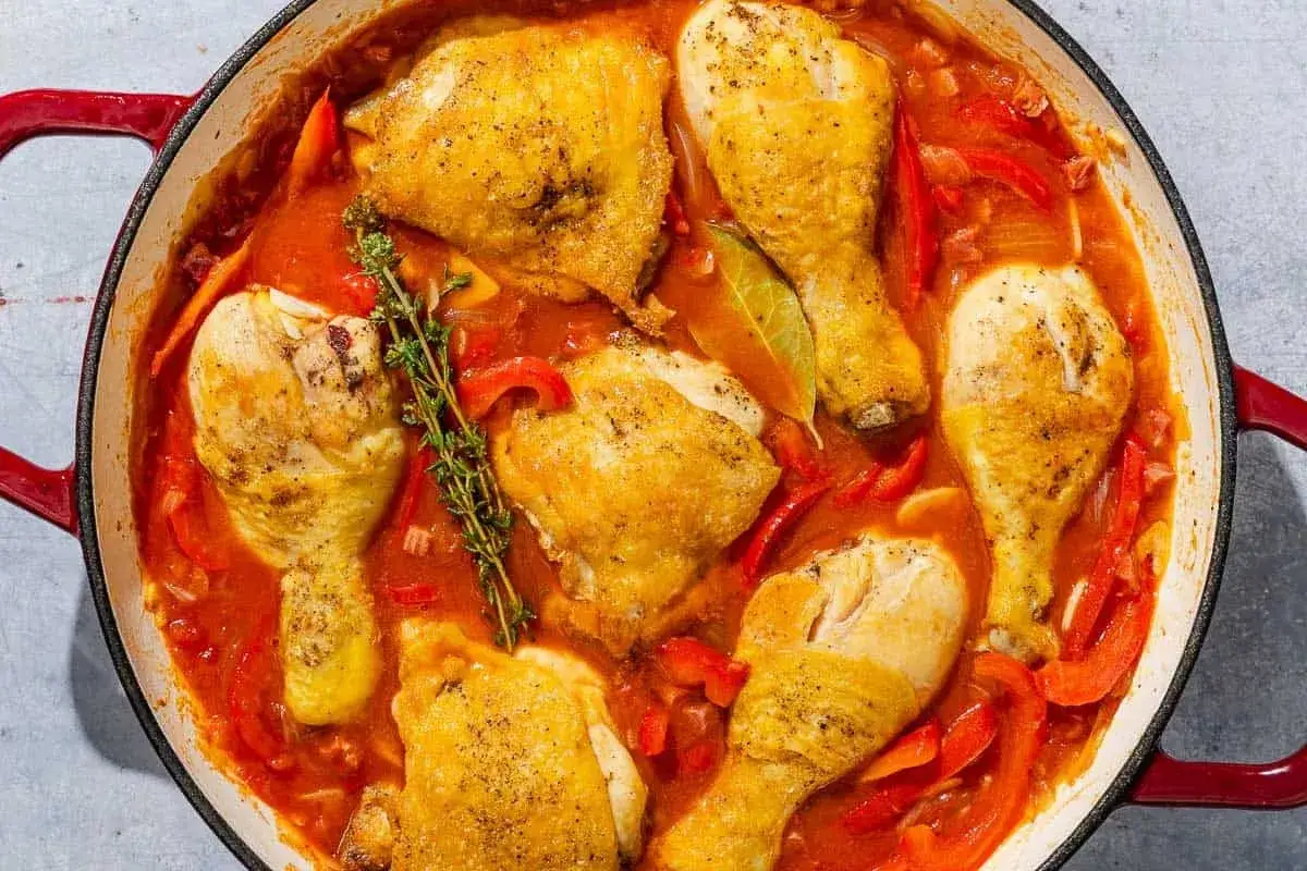 A hearty chicken basquaise simmers in a rich tomato and pepper sauce, garnished with fresh thyme and bay leaves in a red Dutch oven.