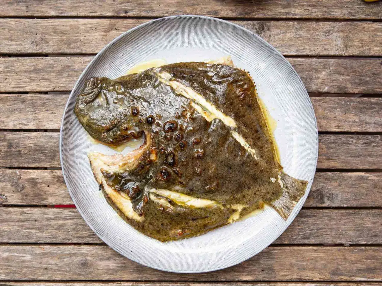 A whole grilled turbot, glistening with butter and herbs, served on a rustic plate atop a wooden table.