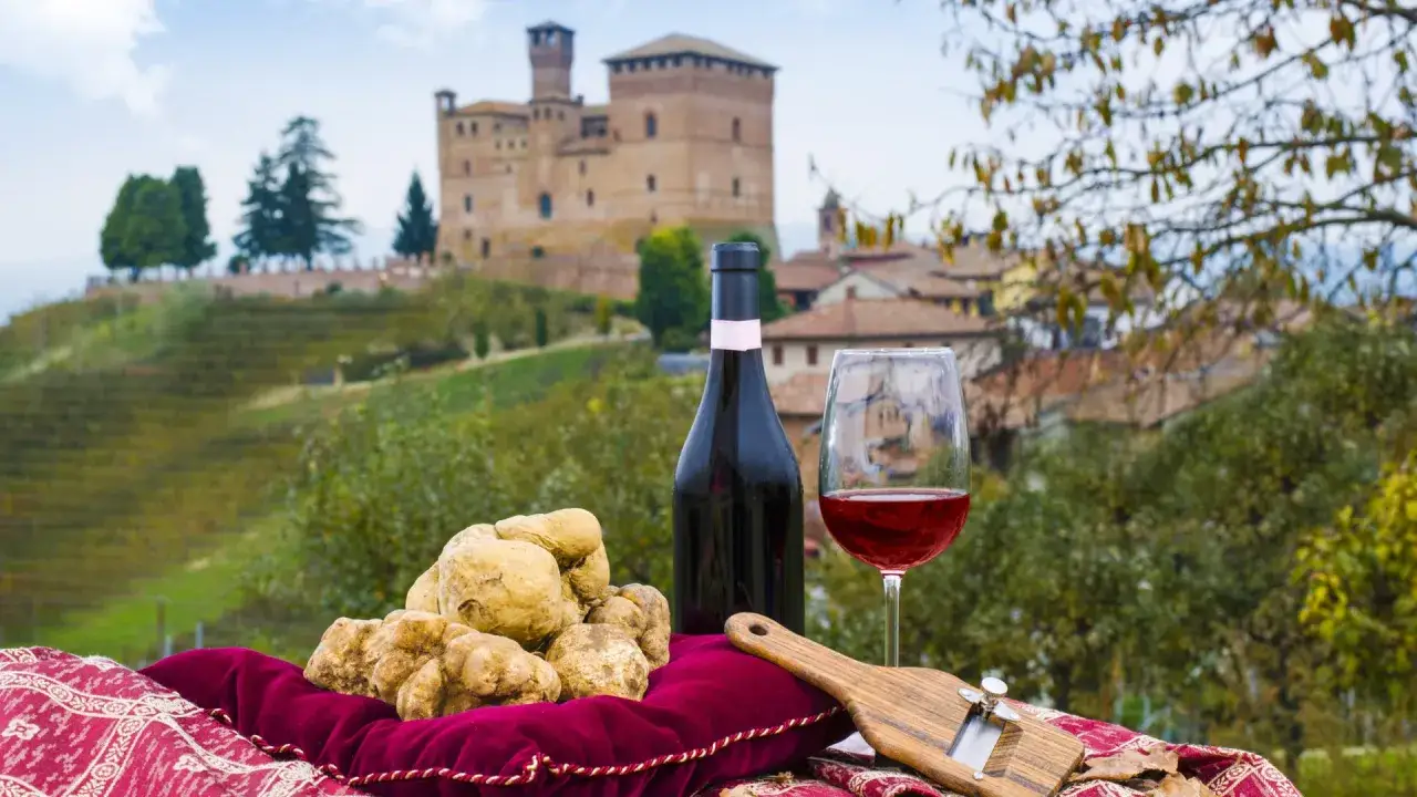 A bottle of red wine, a glass, and fresh truffles are set against a backdrop of rolling hills and a castle in the picturesque Piedmont wine region.